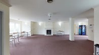 This single-floor space offers a generous, open layout designed for communal gathering and relaxation. The room is anchored by a classic white brick fireplace, serving as a natural focal point and adding warmth to the overall atmosphere. Decorative white columns frame the space, providing architectural definition without interrupting the open flow of the floor plan. Rich, mauve-toned carpeting covers the entire floor, adding texture and a sense of comfort underfoot. The interior is bathed in soft, ambient light from wall-mounted fixtures and ceiling-mounted lighting, contributing to a welcoming and well-lit environment. The neutral white walls and ceiling keep the space feeling bright and clean, complementing the traditional architectural details. Several small tables and chairs are arranged throughout the room, suggesting a multi-purpose use suitable for dining, meetings, or social gatherings. Additional doors visible along the perimeter indicate access to adjoining rooms or utility spaces. The combination of functional furnishings, a central fireplace, and open layout makes this a versatile and inviting shared space.