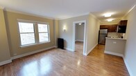 This single-floor residence offers a thoughtfully designed layout with classic architectural details throughout. The living area is characterized by warm, light-toned hardwood floors that extend seamlessly into adjoining spaces, creating a cohesive and inviting feel. Crown molding lines the ceilings, adding a refined touch to the neutral, beige-toned walls that lend the space a clean and timeless aesthetic. A large double-hung window allows natural light to fill the room, enhancing the open and airy atmosphere, while a traditional cast-iron radiator beneath it speaks to the building's classic character. A wall sconce provides warm ambient lighting, complementing the natural light effectively. An open doorway leads to an additional room, also finished with hardwood floors and a ceiling-mounted light fixture, offering versatility in use. To the right, a glimpse of the kitchen reveals stainless steel appliances and dark wood cabinetry, suggesting a functional and well-equipped cooking space. White trim and baseboards throughout provide crisp contrast against the warm flooring and walls, tying the design together cohesively. The overall ambiance is comfortable, classic, and move-in ready.