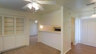 This single-floor space is designed with a practical, open layout that connects the main living areas seamlessly. The kitchen is partially enclosed by a half-wall partition, featuring a pass-through counter that opens toward the living area — a functional detail that allows for easy interaction between spaces while maintaining a degree of separation. Black appliances are visible within the kitchen area, complementing the neutral, light-toned walls throughout.

The living areas are finished with wall-to-wall carpeting in a warm beige tone, contributing to a cozy and understated ambiance. Ceiling fans are installed in multiple rooms, providing both lighting and air circulation. The fan in the primary living space features a decorative white finish, adding a modest ornamental touch.

A doorway visible from the main living area leads into an additional room, also carpeted and equipped with its own ceiling fan, suggesting a versatile secondary space. Built-in shelving is visible along one wall near the entry, offering convenient storage or display options. The overall color palette is neutral and consistent, with white walls and light trim lending the space a clean, simple character throughout.