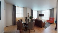 Located at 591 Pearl St in Eugene, Oregon, this thoughtfully designed single-floor unit offers a cohesive blend of modern aesthetics and practical living. The open-plan living and dining area is anchored by large windows dressed with sheer and drape panel combinations, flooding the space with natural light. The living area features a leather sofa, vibrant accent chairs, and a wall-mounted television, while the adjacent dining space includes a round glass-top table with upholstered seating. Warm hardwood-style flooring flows throughout the main areas, complementing the clean white walls and contemporary finishes. The fully equipped kitchen showcases a two-tone cabinetry design in white and dark wood, stainless steel appliances, a subway tile backsplash, and a breakfast bar with teal upholstered seating. Two well-proportioned bedrooms feature plush carpeting, upholstered headboards, ample natural light, and generous closet space. Two bathrooms are finished with dark wood vanities, white quartz countertops, large mirrors, and wood-look tile flooring. A dedicated in-unit laundry closet with a stacked washer and dryer adds everyday convenience. Curated artwork throughout enhances the polished, welcoming ambiance of this residence.