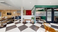 Located at 8650 Kingsbridge Dr in University City, Missouri, this thoughtfully designed single-floor space offers a generous and versatile layout that blends modern aesthetics with eclectic character. The signature bold black-and-white checkerboard tile floor flows throughout, serving as a unifying design element that anchors the entire space.

The expansive main living area is warm and inviting, featuring a white-painted brick fireplace, natural wood accent wall panels, and a large wall-mounted television. Built-in open shelving lines one wall beneath a row of windows that allow abundant natural light to fill the room. Seating areas are thoughtfully arranged to encourage both relaxation and conversation, complemented by recessed lighting overhead.

Toward the rear, a private office enclosure with frosted glass panels and bold teal-green walls provides a focused workspace, clearly branded for professional use. Adjacent to this is a compact kitchenette area featuring navy cabinetry, small appliances, and a charming striped awning detail that adds a playful touch.

The bathroom is a design standout, showcasing dramatic floor-to-ceiling black subway tile, a floating wood-toned vanity, a round brass-framed mirror, and the same checkerboard tile underfoot, tying it cohesively to the rest of the space. Every detail in this space reflects intentional, design-forward curation.