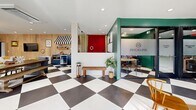 Located at 8650 Kingsbridge Dr in University City, Missouri, this thoughtfully designed single-floor space offers a generous and versatile layout that blends modern aesthetics with eclectic character. The signature bold black-and-white checkerboard tile floor flows throughout, serving as a unifying design element that anchors the entire space.

The expansive main living area is warm and inviting, featuring a white-painted brick fireplace, natural wood accent wall panels, and a large wall-mounted television. Built-in open shelving lines one wall beneath a row of windows that allow abundant natural light to fill the room. Seating areas are thoughtfully arranged to encourage both relaxation and conversation, complemented by recessed lighting overhead.

Toward the rear, a private office enclosure with frosted glass panels and bold teal-green walls provides a focused workspace, clearly branded for professional use. Adjacent to this is a compact kitchenette area featuring navy cabinetry, small appliances, and a charming striped awning detail that adds a playful touch.

The bathroom is a design standout, showcasing dramatic floor-to-ceiling black subway tile, a floating wood-toned vanity, a round brass-framed mirror, and the same checkerboard tile underfoot, tying it cohesively to the rest of the space. Every detail in this space reflects intentional, design-forward curation.