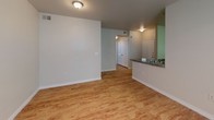 This single-floor residence offers a functional and comfortable layout designed for everyday living. The open living area is finished with light wood-style flooring throughout, creating a warm and cohesive feel across the space. A pass-through counter connects the living area to the kitchen, enhancing the open flow between rooms. The kitchen is well-equipped, featuring warm-toned wooden cabinetry with ample upper and lower storage, granite-style countertops, and a full suite of black appliances including an electric range, over-the-range microwave, and refrigerator. The light flooring in the kitchen complements the cabinetry for a clean, functional aesthetic. The bedroom is generously sized and benefits from natural light through a window, with a ceiling fan providing year-round comfort and light wood flooring continuing the cohesive design. The bathroom is notably elongated, offering a full-length vanity with a single sink, white cabinetry, a large mirror with overhead lighting, a medicine cabinet, and a shower/tub combination with tile surround. Parquet-style flooring adds a classic touch to the bathroom. Overall, the space presents a practical, move-in-ready layout with consistent finishes and thoughtful storage throughout.