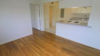 This single-story space features an open-concept layout that connects the main living area to the kitchen, creating a seamless flow throughout. The living area is finished with warm-toned hardwood flooring that adds character and a sense of continuity to the space. A peninsula-style kitchen counter separates the cooking area from the living space, offering both functionality and an open sightline. The kitchen is equipped with white cabinetry and a white range cooktop, lending a clean and classic aesthetic. Transitions from hardwood to light ceramic tile flooring mark the entry into a hallway, which leads to additional rooms accessible through multiple doorways. The walls throughout are painted in a neutral white, contributing to a bright and airy atmosphere. The layout provides a practical flow between the living, cooking, and private areas of the space, making it well-suited for everyday living.