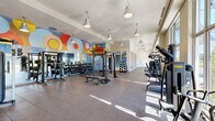 This contemporary fitness center is thoughtfully designed to accommodate a wide range of workout styles and fitness goals. The main gym floor is expansive, featuring rubber flooring throughout and an eye-catching, colorful circular mural that adds a vibrant, energetic atmosphere to the space. Pendant lighting hangs from the ceiling, complementing the natural light that streams in through large windows along the perimeter. The main floor is well-stocked with a comprehensive selection of equipment, including a power rack, cable machines, a dumbbell rack, adjustable benches, a punching bag, kettlebells, medicine balls, and a variety of resistance training machines. A dedicated cardio and strength machine zone runs along one side of the facility, lined with floor-to-ceiling glass partitions that create an open, airy feel while visually connecting the different workout areas. A separate studio room offers a serene environment for yoga, stretching, or dance, featuring smooth hardwood flooring, teal-accented walls, full-length mirrored panels, and a wall-mounted TV. Large windows in this room provide outdoor views, enhancing the calming ambiance. Throughout the facility, the design emphasizes openness and functionality, making it a comprehensive and inviting space for fitness enthusiasts.