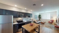 This single-floor residence offers a well-designed open-concept layout that seamlessly connects the kitchen and living area, creating a cohesive and functional living environment. The kitchen is a standout feature, outfitted with dark espresso cabinetry that extends floor to ceiling, providing generous storage. Stainless steel appliances — including a refrigerator, electric range, built-in microwave, and dishwasher — are complemented by crisp white countertops and a single-basin sink. A butcher block kitchen island with bar seating adds both prep space and a casual dining option. Warm hardwood-style flooring runs throughout the space, enhancing the overall sense of continuity and warmth. Recessed lighting in the kitchen and smooth white ceilings keep the interior bright and clean. The living area is generously proportioned, accommodating a full sofa arrangement, accent chair, and media console with ease. A large window allows natural light to fill the room, while neutral walls serve as a versatile backdrop. The space carries a contemporary yet approachable aesthetic, with a comfortable ambiance well-suited for both everyday living and entertaining.