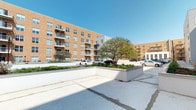 This contemporary residential building presents a striking multi-story brick exterior with a warm, earthy tone that gives it a welcoming urban character. The façade is punctuated by large windows that allow abundant natural light into each unit, while private balconies extend outward from multiple floors, offering residents their own outdoor retreats with open views of the surrounding area. The building's design blends modern architectural lines with classic brick construction, creating a balanced aesthetic that feels both refined and approachable. At ground level, the property features well-maintained landscaped planters with lush greenery and mature trees, adding a natural softness to the hardscape surroundings. The outdoor common areas are finished with clean concrete surfaces that provide ample open space. A secondary wing of the building, clad in a lighter exterior finish, provides architectural contrast and visual interest to the overall composition. The property benefits from clear sightlines and an open sky above, enhancing the sense of space and airiness throughout the exterior. Parking is conveniently accessible on the grounds, contributing to the practical, resident-focused design of this well-appointed building.