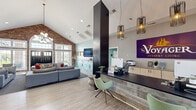 Located at 3535 E Tenth St in Greenville, NC, this single-story leasing and amenity center for The Vantage Studios makes a striking first impression. The grand entrance features soaring vaulted ceilings with an arched transom window above bold black double doors, flooding the space with natural light and creating an airy, welcoming atmosphere. Light wood-look flooring flows throughout, complemented by a mix of exposed brick, soft teal and white walls, and a dramatic floor-to-ceiling dark column anchoring the reception area. The front desk, clad in reclaimed wood-style paneling, is outfitted with modern pendant lighting and stylish seating, establishing a polished yet approachable tone. The expansive lounge area features generous gray sectional sofas, colorful accent pillows, and contemporary light fixtures, ideal for relaxed social gatherings. A cozy side lounge offers leather and upholstered chairs arranged around a round coffee table, accented by bold teal wallcovering and yellow industrial sconces. A media room with a dramatic dark accent wall adorned with large typographic lettering, a flat-screen TV, and soft globe lighting provides an intimate entertainment retreat. A vending and dining area rounds out the space, offering a communal table with vibrant red seating alongside snack and beverage machines, with direct access to the outdoors.
