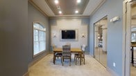 This single-floor space exudes a refined and welcoming ambiance, thoughtfully designed with a blend of classic architectural details and modern touches. The main room features a distinctive tray ceiling with recessed lighting, adding depth and an elevated sense of height to the space. Soft blue-gray walls are complemented by crisp white crown molding and decorative trim, creating a polished, cohesive aesthetic throughout.

A focal point of the room is a framed wall niche housing a wall-mounted flat-screen television, flanked by stylish wall sconces that provide warm, ambient lighting. An arched window dressed with plantation shutters allows natural light to filter in, brightening the neutral-toned carpeted floor. The seating area centers around a wooden dining table surrounded by plaid upholstered chairs, lending a comfortable yet elegant feel suited for meetings or small gatherings.

A large mirror panel along one wall enhances the sense of openness, visually expanding the space while offering a glimpse into an adjoining area. The overall design strikes a balance between functionality and sophistication, making it a versatile and inviting environment for a variety of uses.