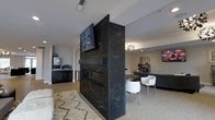 Located at 1 Mirada Dr N in Lewis Center, Ohio, this single-floor space offers a contemporary and polished environment designed for comfort and functionality. The open-concept layout is anchored by a striking double-sided linear fireplace clad in dark marble-veined stone, which serves as a dramatic architectural divider between two distinct lounge zones. A wall-mounted flat-screen television above the fireplace enhances the space's modern aesthetic.

On one side of the fireplace, a cozy seating area features a plush area rug and soft furnishings, creating an inviting retreat. On the opposite side, a casual dining or social zone is set with a round dark table and sleek white chairs, illuminated by an elegant chandelier overhead. A large sectional sofa and an additional mounted display screen define a third relaxation area, ideal for gathering.

A dedicated refreshment station with cabinetry, counter space, and appliances is tucked along one wall, adding a practical amenity to the space. Throughout, light wood-toned flooring, neutral wall tones, and recessed lighting contribute to a clean, upscale atmosphere. The cohesive design blends sophisticated materials with functional layout, making this space well-suited for community use, hospitality, or professional entertaining.