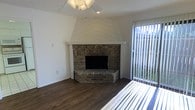This two-story space combines classic design elements with practical functionality. The main living area immediately draws attention with its standout corner stone fireplace, framed by a striking white chevron-patterned mantel surround that extends to the ceiling, creating a strong architectural focal point. The hearth is finished in matching stacked stone, adding to the rustic character of the room.

The flooring throughout features rich, dark wood-look planks that provide warmth and contrast against the crisp white walls. A ceiling fan is centrally positioned, ensuring year-round air circulation and comfort. Natural light enters through vertical blind-covered windows, offering both brightness and privacy control.

The textured ceiling adds a layer of visual depth typical of residential construction, while the clean white walls provide a neutral canvas adaptable to a variety of interior styles. The layout spans two floors, offering separation of living and private spaces. Overall, the space carries a warm, inviting ambiance rooted in its traditional architectural details, particularly the prominent fireplace, which serves as the heart of the main living area.