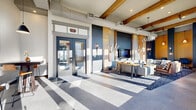 Located at 300 Enterprise Blvd in Bozeman, Montana, this single-floor lobby lounge is a thoughtfully designed communal space that blends modern aesthetics with warm, natural materials. Soaring ceilings with exposed wood beams run the length of the space, creating an open, airy atmosphere that is further enhanced by an expansive wall of floor-to-ceiling windows. These windows flood the interior with abundant natural light and offer clear views of the surrounding Bozeman landscape.

The space is organized into two distinct zones. A comfortable lounge area anchors one end, featuring a bold navy blue feature wall adorned with wall-mounted media and flanked by warm wood paneling. Stylish seating — including white sofas, leather accent chairs, and a patterned area rug — centers around a wood-topped coffee table beneath a geometric chandelier, creating an inviting gathering space.

Toward the entry, a casual dining area features sturdy wood high-top tables paired with leather barstools, illuminated by sleek black pendant lights. Large-format tile flooring flows throughout, transitioning seamlessly between zones. The overall design is contemporary and refined, balancing industrial elements like exposed ductwork with the warmth of natural wood and rich, curated furnishings.
