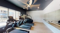 This single-level fitness center is designed with both functionality and aesthetics in mind. The space features a striking design contrast, with dark navy-painted ceilings and accent walls paired against crisp white lower walls, creating a modern and energizing atmosphere. Light wood-tone flooring runs throughout, providing a clean, polished foundation underfoot.

The facility is outfitted with a lineup of TRUE-brand cardio equipment, including multiple treadmills and stair climbers, all arranged to maximize the use of the available floor space. A wall-mounted mirror along one side of the room enhances the sense of openness and allows users to monitor their form during workouts.

Large sliding glass windows and doors along one wall flood the interior with natural light and offer direct views of an outdoor pool and lounge area, making for an inspiring workout environment. A ceiling fan overhead provides additional airflow and comfort during exercise sessions.

Practical amenities include a hand sanitizer station, a waste receptacle near the entry door, and clearly marked exit signage. The entry door features a glass panel, maintaining visual connectivity with adjacent spaces. Recessed lighting supplements the natural light for well-distributed illumination throughout the room. Overall, this fitness center delivers a clean, contemporary space tailored for effective and comfortable workouts.