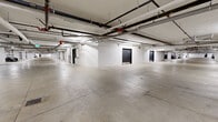 This underground parking facility encompasses a generously sized single-level layout, providing ample space for vehicle and bicycle storage. The floor plan follows a cross-shaped configuration, maximizing the usable area and allowing for efficient traffic flow throughout the garage. The concrete floor is clearly marked with numbered stalls and painted lane lines, ensuring organized and orderly parking. Designated accessible parking spaces are also present, highlighting the facility's inclusive design. The ceiling features an exposed infrastructure system, including a network of plumbing pipes, ventilation ducts, fire suppression lines, and fluorescent lighting fixtures that illuminate the space evenly. Support columns are positioned throughout, complementing the structural integrity of the overhead system. Mechanical equipment, including ventilation units, is integrated into select areas of the garage. Several dark-painted utility doors provide access to adjoining spaces or service areas. Dedicated bicycle parking is available, with bike racks accommodating multiple bicycles in designated zones. The white-painted walls and well-lit environment contribute to a clean and functional atmosphere. Overall, this facility is well-equipped to serve the parking and storage needs of a multi-unit residential or commercial building.