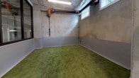 This single-floor pet care space is designed with both functionality and durability in mind, showcasing an industrial aesthetic rooted in exposed cast concrete walls and polished concrete floors throughout. The main area is open and spacious, offering ample room for pet grooming and handling. A professional-grade stainless steel grooming tub with an integrated step and utility connections anchors one end of the space, supported by visible copper plumbing lines and dedicated water supply fixtures along the walls — all purposefully left exposed for easy access and maintenance.

Overhead, open-ceiling infrastructure reveals ductwork, insulated HVAC components, and fluorescent strip lighting that provides bright, even illumination across the entire floor. A large bank of dark-framed windows runs along one interior wall, offering views into an adjacent area and allowing natural light to filter in, lending an open and connected feel to the space.

A separate room features artificial turf flooring — a practical and hygienic relief zone for animals — with coated lower wall panels for easy cleaning and small clerestory windows that introduce additional natural light. Exposed utility pipes and drainage infrastructure in this zone further underscore the space's purpose-built, functional design. Together, the areas form a well-considered layout tailored for professional pet grooming or animal care operations.