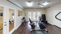 This fitness center offers a thoughtfully organized workout environment across a single floor, designed to accommodate a range of exercise needs. The space features dark hardwood-style flooring throughout, providing a clean and durable surface well-suited for active use. Neutral beige walls and bright overhead fluorescent lighting, supplemented by ceiling fans, create a comfortable and well-ventilated atmosphere.

The cardio area is outfitted with multiple True-brand machines, including treadmills, ellipticals, and stationary and recumbent bikes, each positioned on protective floor mats. A wall-mounted flat-screen television is positioned for easy viewing during workouts. A dedicated free-weight station includes a full dumbbell rack with varying weight increments.

The space is divided into connected zones, with interior glass-panel French doors offering views between sections and allowing natural light to flow through. Large exterior French doors open toward an outdoor pool area, enhancing the sense of openness. Colorful framed artwork adds visual interest to the walls. Additional features include a water station and adjacent access to a kitchenette area, making this fitness center a comprehensive and convenient workout destination.