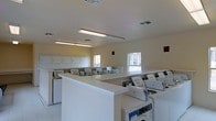 This single-floor communal laundry facility is designed with practicality and efficiency in mind. The room is outfitted with rows of top-loading washing machines and dryers arranged along the perimeter and central areas, providing ample capacity for multiple users simultaneously. Wide, white-finished folding counters run the length of the room, offering generous surface space for sorting and folding laundry. Overhead, fluorescent strip lighting ensures the space is brightly and evenly illuminated throughout. The walls are finished in a neutral cream tone, contributing to a clean and orderly atmosphere. Ventilation is maintained through ceiling-mounted HVAC vents, keeping the environment comfortable. A window along one wall allows for natural light to supplement the artificial lighting, offering a view to the exterior. Upper cabinetry lines one wall, providing additional storage for residents. The light-colored tile flooring is easy to maintain and reinforces the facility's hygienic, no-fuss character. Overall, this laundry room is a well-organized, fully functional shared amenity space built to serve the practical needs of its users efficiently.