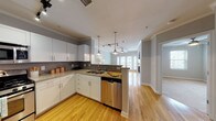 This single-floor residence combines modern finishes with a functional, open layout. The kitchen serves as a central highlight, showcasing white shaker-style cabinetry, granite countertops with a subtle speckled pattern, and a tasteful gray tile backsplash. A peninsula with bar-height seating separates the kitchen from the living area, while pendant lighting and a track light system above add warmth and character. Stainless steel appliances — including a gas range, microwave, refrigerator, and dishwasher — complete the well-equipped cooking space. A stackable in-unit washer and dryer is conveniently tucked just off the kitchen. Warm hardwood flooring flows throughout the main living areas, complemented by crown molding and a ceiling fan that enhance the open feel. The bedroom is finished with plush carpeting, a ceiling fan with integrated lighting, and a window with blinds that brings in natural light. The bathroom features neutral ceramic tile flooring and a tub-shower combination, maintaining a clean and classic aesthetic. Throughout the space, soft gray walls and white trim create a cohesive, modern palette. French doors at the end of the living area invite natural light and offer an outdoor connection.