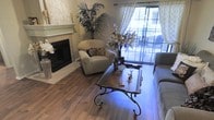 This single-story space centers around a well-appointed living room that blends classic and contemporary design elements. The room features warm hardwood-style flooring that anchors the space, complemented by a white-mantled fireplace with tile surround that serves as a focal point along one wall. Sheer patterned curtains frame sliding glass doors, allowing natural light to filter into the room while providing a seamless connection to an outdoor patio area, where additional seating is visible beyond the glass.

The living area is furnished with plush, neutral-toned seating, including a rounded armchair and a sofa, arranged around an ornate wrought-iron and wood coffee table that adds a decorative, old-world character to the space. Decorative accents, including a tall indoor palm, floral arrangements, and sculptural vases, lend a layered, curated aesthetic throughout. The combination of soft textiles, natural greenery, and warm flooring tones creates an inviting and comfortable ambiance, making the living area both functional and visually rich.