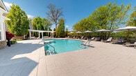 Located at 116 Danbury Rd in Wilton, Connecticut, this outdoor aquatic amenity area offers a well-appointed resort-style pool experience set against a backdrop of mature, lush trees. The centerpiece is a generously sized freeform swimming pool with crystal-clear turquoise water, accessible via stainless steel handrail entry points and featuring a gradual shallow end. Adjacent to the main pool is a separate circular wading pool, ideal for leisure or cooling off. The expansive pool deck is finished in neutral-toned pavers, providing ample space for relaxation and movement. A row of cushioned chaise lounge chairs, each paired with a white market umbrella, lines the perimeter, offering comfortable shaded seating throughout the day. A white pergola structure at one end of the deck adds an architectural focal point and additional shaded retreat space. The area is fully enclosed by a sleek fence, ensuring privacy and security. A stone accent wall and a clubhouse-style building with classic architectural detailing are visible at the pool's edge, adding character to the space. The surrounding canopy of deciduous and evergreen trees creates a serene, park-like atmosphere, making this outdoor space a tranquil and inviting destination.