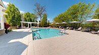 Located at 116 Danbury Rd in Wilton, Connecticut, this outdoor aquatic amenity area offers a well-appointed resort-style pool experience set against a backdrop of mature, lush trees. The centerpiece is a generously sized freeform swimming pool with crystal-clear turquoise water, accessible via stainless steel handrail entry points and featuring a gradual shallow end. Adjacent to the main pool is a separate circular wading pool, ideal for leisure or cooling off. The expansive pool deck is finished in neutral-toned pavers, providing ample space for relaxation and movement. A row of cushioned chaise lounge chairs, each paired with a white market umbrella, lines the perimeter, offering comfortable shaded seating throughout the day. A white pergola structure at one end of the deck adds an architectural focal point and additional shaded retreat space. The area is fully enclosed by a sleek fence, ensuring privacy and security. A stone accent wall and a clubhouse-style building with classic architectural detailing are visible at the pool's edge, adding character to the space. The surrounding canopy of deciduous and evergreen trees creates a serene, park-like atmosphere, making this outdoor space a tranquil and inviting destination.