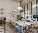 This expansive 2,380 square foot single-story community space in Corinth, TX, showcases a harmonious blend of modern design and functional amenities. The interior boasts high ceilings adorned with striking light fixtures, including a spectacular sunburst chandelier. Wood-look tile flooring runs throughout, accented by inlaid geometric patterns that add visual interest.

The main lounge area features plush seating in soothing blue tones, complemented by accent chairs and metallic side tables. A focal point is the sleek entertainment center with a large TV mounted on a marble-look backdrop, flanked by illuminated display niches. Adjacent to this, a gourmet kitchen with stainless steel appliances and a generous island with bar seating provides an ideal space for casual dining and social gatherings.

Other amenities include a game room equipped with arcade machines and a pool table, perfect for entertainment. Large windows and glass doors allow natural light to flood the space, while offering views to the outdoor area. Thoughtful design elements such as arched doorways, accent walls in calming blue hues, and curated wall art contribute to the sophisticated ambiance. This versatile space is designed to cater to various social activities and relaxation needs of community members.