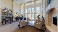 This elegantly appointed clubhouse offers a grand, open layout defined by dramatic double-height ceilings and stately white columns that lend a sense of architectural grandeur throughout the space. The main lounge area centers around a striking stone-surround fireplace, flanked by gold-accented credenzas and framed artwork against a teal accent wall, creating a warm and inviting focal point. Comfortable herringbone-upholstered armchairs and a marble-top coffee table are thoughtfully arranged to encourage conversation and relaxation. A wall of tall, arched, floor-to-ceiling windows floods the interior with abundant natural light while offering views of the outdoor pool and landscaped grounds beyond. Soft, neutral tones in the carpeting and walls are accented by vibrant touches of cobalt blue velvet seating, lush tropical plantings, and decorative woven wall art, adding visual interest and texture. A separate bar-height seating area with counter stools and open shelving provides a casual gathering spot, while French doors lead to additional rooms within the facility. The overall ambiance is polished yet welcoming, blending classic architectural details with contemporary furnishings to create a versatile community space ideal for both social gatherings and quiet retreats.