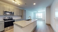 This single-floor unit offers a clean, contemporary design highlighted by light wood-look flooring, crisp white walls, and modern finishes throughout. The open-concept layout seamlessly connects the kitchen and living area, creating a bright and functional shared space. The kitchen is a standout feature, equipped with granite countertops, a central island with an undermount sink and brass-toned faucet, gray shaker-style cabinetry, and a full suite of stainless steel appliances including a refrigerator with ice maker, electric range, built-in microwave, and dishwasher. Track lighting and recessed fixtures illuminate the kitchen, while the adjacent living area benefits from natural light streaming through large windows fitted with horizontal blinds. A ceiling fan with integrated lighting is present in both the living area and bedroom, enhancing comfort and airflow. The bedroom is generously sized with an open, airy feel and direct access to the en-suite bathroom. The bathroom features a dual-sink vanity topped with granite countertops, gray cabinetry, framed mirrors, and a tall storage cabinet, offering both style and practicality. The overall ambiance of the space is modern and move-in ready, with a neutral palette that provides a versatile backdrop for any décor style.