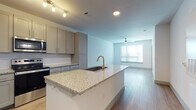 This single-floor unit offers a clean, contemporary design highlighted by light wood-look flooring, crisp white walls, and modern finishes throughout. The open-concept layout seamlessly connects the kitchen and living area, creating a bright and functional shared space. The kitchen is a standout feature, equipped with granite countertops, a central island with an undermount sink and brass-toned faucet, gray shaker-style cabinetry, and a full suite of stainless steel appliances including a refrigerator with ice maker, electric range, built-in microwave, and dishwasher. Track lighting and recessed fixtures illuminate the kitchen, while the adjacent living area benefits from natural light streaming through large windows fitted with horizontal blinds. A ceiling fan with integrated lighting is present in both the living area and bedroom, enhancing comfort and airflow. The bedroom is generously sized with an open, airy feel and direct access to the en-suite bathroom. The bathroom features a dual-sink vanity topped with granite countertops, gray cabinetry, framed mirrors, and a tall storage cabinet, offering both style and practicality. The overall ambiance of the space is modern and move-in ready, with a neutral palette that provides a versatile backdrop for any décor style.