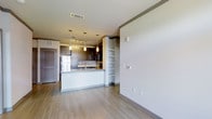 This single-floor space showcases a thoughtfully designed open-concept layout that seamlessly connects the kitchen and living areas. The kitchen serves as a focal point, featuring a sleek white peninsula with a stone countertop, dark espresso cabinetry, and a full suite of stainless steel appliances including a refrigerator and microwave. A trio of pendant lights hangs above the peninsula, providing both task lighting and a modern aesthetic touch, while a track lighting system illuminates the kitchen workspace. A classic tile backsplash adds texture and visual interest behind the cabinetry. Built-in open shelving provides both practical storage and display space, bridging the kitchen to the adjoining living area. Light-toned hardwood flooring flows continuously throughout the space, contributing to an airy, cohesive feel. The neutral color palette of soft whites and warm grays creates a clean, contemporary ambiance. A louvered door adds a subtle architectural detail, while recessed HVAC vents blend seamlessly into the ceiling. The open layout allows for flexible furniture arrangements and maximizes the sense of space throughout this well-appointed residence.