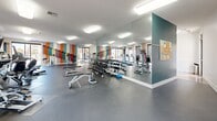 Located at South Junction in Memphis, Tennessee, this single-floor fitness center is designed to support a comprehensive workout experience. The space is visually distinctive, featuring bold geometric accent walls in a striking palette of orange, teal, lavender, and muted yellow — creating an energetic atmosphere that sets the tone for exercise.

The facility is generously proportioned, providing ample room to accommodate a wide variety of equipment without feeling crowded. Cardio equipment includes treadmills, elliptical machines, and stationary bikes, all positioned near large windows and sliding glass doors that allow natural light to fill the space and offer views of the surrounding neighborhood. A wall-mounted flat-screen television adds entertainment options for cardio sessions.

The strength training area features a multi-functional cable machine, adjustable benches, and a full rack of dumbbells. Floor-to-ceiling mirrors line one wall, enhancing the sense of openness and allowing users to monitor their form throughout workouts. Rubber flooring throughout provides safety and durability.

Flush-mounted ceiling lights ensure consistent, bright illumination across the entire floor, while roller shades on all windows offer adjustable light control. A bulletin board near the entrance provides a functional touch for community notices.