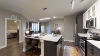This single-floor residence offers a thoughtfully designed open-concept layout that seamlessly connects the kitchen, dining, and living areas. The kitchen serves as the centerpiece of the space, anchored by a large island with a crisp white quartz countertop, an undermount sink, and a built-in dishwasher. Dark wood cabinetry on the island provides a striking contrast to the lighter finishes throughout. Bar-height seating along the island creates an inviting spot for casual dining and entertaining. Stainless steel appliances, including a full-size refrigerator, complete the modern kitchen aesthetic. Track lighting and recessed ceiling fixtures ensure the space is well-lit throughout. Warm hardwood-style flooring extends across the open floor plan, adding a sense of continuity and warmth. The living area, visible beyond the kitchen, features a ceiling fan and access to natural light through large windows dressed with curtains. Neutral gray walls provide a versatile backdrop that complements the contemporary furnishings and décor. The overall ambiance is clean, modern, and functional, making this space well-suited for both everyday living and entertaining guests.