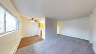 This single-floor residence offers a practical and comfortable layout designed for everyday living. The open living area is finished with plush gray carpeting, creating a warm and inviting atmosphere, while the adjoining space transitions to smooth hardwood-style flooring for a clean contrast. A ceiling fan with an integrated light fixture is centrally mounted, providing both airflow and ambient lighting to the main living area. A partial wall divider separates the living space from the kitchen, lending a sense of definition without fully closing off the floor plan. The kitchen features warm, honey-toned wood cabinetry that adds a classic character to the space, complemented by under-cabinet lighting that brightens the work area. A baseboard heater is visible along the wall, ensuring efficient climate control throughout the unit. The neutral, light-toned walls and textured ceiling provide a clean, understated backdrop that makes the space feel bright and easy to personalize. The overall layout is efficient and functional, offering a cohesive living environment with a straightforward, welcoming design that suits a variety of lifestyles.