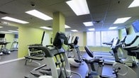 This fitness center offers a comprehensive workout environment designed to accommodate a wide range of exercise needs. The space is outfitted with an array of cardio machines, including ellipticals, stationary bikes, and treadmills, all arranged to allow comfortable movement between stations. A dedicated free weights area features a full dumbbell rack, providing options for strength and resistance training. The interior is finished with light green accent walls and structural columns that add a touch of color to the otherwise clean and neutral aesthetic. A dropped ceiling with recessed panel lighting ensures the space is evenly and brightly lit throughout. Large windows fitted with vertical blinds allow natural light to filter in, contributing to an open and energizing atmosphere. Full-length mirrors are positioned along the walls, enhancing the sense of space while allowing users to monitor their form during workouts. The flooring is finished in a dark, durable material suited for high-traffic fitness use. Overall, the facility presents a practical, well-maintained environment that supports both cardiovascular and strength training goals in a comfortable and organized setting.