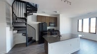 This two-story space combines contemporary design with functional living. The kitchen serves as a central feature, outfitted with dark-toned cabinetry, black appliances including a refrigerator, range, and microwave, and a peninsula with a dark countertop and integrated sink. Track lighting overhead provides focused illumination throughout the cooking area. The clean white walls contrast with the rich wood-toned cabinetry and dark hardwood-style flooring, creating a modern, cohesive aesthetic. A standout architectural element is the black metal spiral staircase positioned adjacent to the kitchen, connecting the main level to the upper floor. Its compact, sculptural design adds a bold visual focal point while maximizing the use of floor space. The open layout of the main level promotes a seamless flow between areas, contributing to an airy, uncluttered atmosphere. The combination of dark finishes, crisp white walls, and industrial-style details gives this space a distinctly contemporary character suited to modern living.