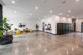 This contemporary building lobby makes a strong first impression with its clean, minimalist design and open layout. Polished concrete floors extend throughout the entire space, reflecting recessed overhead lighting and contributing to a bright, airy atmosphere. The grand entry features sleek double glass doors flanked by floor-to-ceiling windows, allowing natural light to flood the interior, and is accented by two large geometric planters housing lush fiddle-leaf fig trees.

The lounge area is thoughtfully designed with a neutral sectional sofa and matching tufted couch, accented by bold yellow cube ottomans, a sculptural wooden coffee table, and a patterned area rug. A large abstract artwork anchors the lounge wall, adding a curated, artistic touch. Tall indoor plants are strategically placed throughout to bring warmth and life to the space.

Adjacent to the lounge, a dedicated mailroom area houses an extensive bank of dark metal resident mailboxes, providing a secure and organized mail solution. A stainless steel elevator with clearly posted building signage offers convenient vertical access. Further into the lobby, a wall-mounted flat-screen TV and additional seating areas, including a dining space with colorful pendant lighting visible through the windows, enhance the functionality and social character of this welcoming communal space.