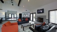 This single-story space functions as a stylish communal lounge, distinguished by its soaring vaulted ceilings accented with dark exposed beams that add architectural character to the bright, open interior. The high ceilings amplify the sense of spaciousness throughout the room, while recessed lighting provides consistent illumination across the space.

The seating area is anchored by sleek black leather sofas and chairs accented with bold orange throw pillows, arranged around a marble-topped coffee table and a plush area rug. A wall-mounted flat-screen television serves as a central entertainment feature, positioned above a classic fireplace framed in a clean, modern surround with a glass-paneled screen — a notable focal point of the room.

Along the back wall, a wet bar or kitchenette area is visible, equipped with a countertop, cabinetry, and dual framed mirrors, providing functional amenities for gatherings. French doors adjacent to the fireplace allow natural light to filter into the space while offering direct access to an outdoor patio or courtyard beyond. The overall design balances contemporary style with comfortable functionality, making it well-suited for social gatherings or relaxed lounging.