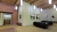 This single-floor space masterfully combines historic industrial character with modern interior design sensibilities. The most striking architectural feature is the soaring ceiling, where substantial exposed wooden beams span the width of the room, lending a raw, structural elegance to the space. Complementing this are the original exposed brick walls, which add warmth, texture, and a sense of history to the interior.

The flooring is covered in a detailed, patterned encaustic-style tile in muted greens and neutrals, providing a sophisticated visual anchor that ties the space together. Pendant lighting fixtures hang at varying heights from the ceiling, providing a clean, modern contrast to the rustic overhead structure and ensuring the space is well-lit throughout.

The main area functions as a gallery-like lounge, with smooth, light green-painted walls serving as a refined backdrop for a curated selection of framed black-and-white landscape photographs. A smaller framed green artwork adds a subtle color accent. Seating includes a sleek black leather sofa and a simple wooden bench, balancing comfort with minimalism. A white-framed door on one wall suggests additional spaces beyond. The overall ambiance is calm and contemplative, blending industrial heritage with artistic refinement.