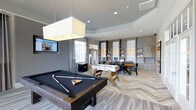 This single-floor amenity space is thoughtfully designed to serve as both a recreation hub and a functional gathering area. The layout is open and expansive, anchored by a sleek billiards table positioned beneath a large, square pendant light that provides focused illumination. A foosball table sits nearby, adding to the game room atmosphere. The flooring throughout features a distinctive chevron-patterned hardwood in warm, neutral tones that contrasts beautifully with the cool grey walls and tray ceilings, lending the space a contemporary yet inviting feel. Recessed lighting and pendant fixtures illuminate the room evenly, complementing the natural light that filters in through large windows dressed with floor-length curtains. Along the far wall, a built-in shelving and cabinetry unit in light wood tones houses workstations equipped with desktop computers, making this space equally suited for productivity. Dining-height seating and a table are positioned nearby for collaborative use. A wall-mounted flat-screen television adds to the entertainment offerings. The overall ambiance is modern and polished, balancing recreational amenities with workspace functionality in a well-appointed, multipurpose environment.