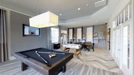 This single-floor amenity space is thoughtfully designed to serve as both a recreation hub and a functional gathering area. The layout is open and expansive, anchored by a sleek billiards table positioned beneath a large, square pendant light that provides focused illumination. A foosball table sits nearby, adding to the game room atmosphere. The flooring throughout features a distinctive chevron-patterned hardwood in warm, neutral tones that contrasts beautifully with the cool grey walls and tray ceilings, lending the space a contemporary yet inviting feel. Recessed lighting and pendant fixtures illuminate the room evenly, complementing the natural light that filters in through large windows dressed with floor-length curtains. Along the far wall, a built-in shelving and cabinetry unit in light wood tones houses workstations equipped with desktop computers, making this space equally suited for productivity. Dining-height seating and a table are positioned nearby for collaborative use. A wall-mounted flat-screen television adds to the entertainment offerings. The overall ambiance is modern and polished, balancing recreational amenities with workspace functionality in a well-appointed, multipurpose environment.