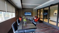 This single-floor space functions as a stylish multipurpose lounge or amenity room, combining a formal meeting or dining area with a comfortable social seating zone. The main area features a long, dark glass-topped table flanked by contemporary black chairs, ideal for gatherings or meetings. Linear LED lighting strips run across the ceiling, casting a clean, modern glow over the space.

A wall-mounted flat-screen television is positioned prominently on a textured accent wall, serving both the dining and lounge areas. The adjacent seating zone includes bold red upholstered chairs arranged around a small round side table, adding a vibrant contrast to the otherwise neutral, dark color palette of grays and blacks.

A large glass partition separates the main lounge from an adjoining room, enhancing the sense of depth while maintaining visual connectivity. Framed artwork decorates the walls, contributing a polished, curated ambiance. Patterned carpeting with red and gray tones grounds the space and ties the color scheme together cohesively. A small decorative plant centerpiece on the table introduces a natural element to the otherwise sleek, urban interior design.