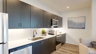 This thoughtfully designed one-bedroom, one-bathroom residence offers a clean, modern aesthetic throughout its efficiently utilized single-floor layout. Light wood-style flooring flows seamlessly through the main living areas, complementing the bright white walls and recessed lighting that keep the space feeling open and airy.

The kitchen is a standout feature, showcasing dark charcoal cabinetry with brushed gold hardware, white quartz countertops, a subway tile backsplash, and a full suite of stainless steel appliances including a range, microwave, dishwasher, and refrigerator. The layout provides ample counter and storage space for everyday cooking.

The living room is generously proportioned, with a sliding glass door that opens to a private balcony overlooking the surrounding urban landscape, allowing abundant natural light to fill the space. The open flow between the living area and kitchen makes the layout feel expansive and functional.

The bedroom offers a comfortable and well-lit retreat, featuring plush carpet, a large window with outdoor views, and enough space for a full furniture arrangement. The bathroom is finished with a wide quartz vanity, dark cabinetry with gold accents, and a sleek LED-backlit mirror, echoing the refined design language found throughout the home.