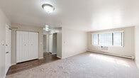 This single-floor unit is designed with a straightforward, functional layout that makes efficient use of its space. The living area is generously sized, finished with wall-to-wall neutral carpet and bright white walls that contribute to an open, airy feel. A large window fitted with vertical blinds allows natural light to fill the room, while a baseboard heating system and wall-mounted AC unit provide year-round climate control. Flush-mount ceiling fixtures provide consistent overhead lighting throughout.

The kitchen features wood-look vinyl plank flooring, white cabinetry, a gas range, and a full-size black refrigerator. A hallway connects the kitchen to the rest of the unit, maintaining a clear flow between living and private spaces.

The bedrooms are carpeted and well-proportioned, each offering closet space. One bedroom includes an adjacent walk-in closet with a rod and shelving system, while natural light enters through windows fitted with baseboard heaters beneath.

The bathroom includes a full tub-shower combination with white tile surround, a curved shower rod, brushed nickel fixtures and hardware, and neutral-toned walls. Throughout the unit, the consistent use of light, neutral tones and clean finishes creates a calm, move-in-ready environment.