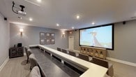 Located at 27821 Alabama 181 in Daphne, Alabama, this single-floor space is designed as a dedicated screening and presentation room that combines functionality with refined aesthetics. The centerpiece of the room is a large projection screen mounted at the front, supported by a decorative ornate media console that adds a touch of classic elegance to the space. Rows of upholstered gray chairs are arranged in a theater-style layout, complemented by long white ledge tables that run the length of each row, providing a practical surface for guests. The flooring features a dark concrete finish that grounds the room with a modern, polished look. Soft gray walls create a neutral, cinema-like ambiance, while recessed ceiling lights and warm wall sconces offer layered lighting suited for both presentations and screenings. A curated gallery of framed photographs adorns one wall, adding character and visual interest to the space. A white-trimmed interior door provides access, and the overall design strikes a balance between a professional presentation environment and a comfortable, stylish gathering space. The room's thoughtful layout and upscale finishes make it well-suited for private screenings, corporate events, or community gatherings.