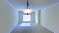 This single-floor space presents a clean, well-maintained interior highlighted by a spacious main room visible in the image. The room features soft, neutral-toned wall-to-wall carpeting that contributes to a calm and comfortable atmosphere. Crown molding lines the upper perimeter of the walls, adding a subtle architectural detail that elevates the overall finish of the space.

A prominent bowl-style pendant light fixture with a dark bronze frame hangs centrally from the ceiling, serving as a functional and decorative focal point. The ceiling height appears standard, providing an open and airy feel throughout the room.

Natural light enters generously through two windows fitted with white horizontal blinds, casting soft patches of sunlight onto the carpeted floor. A door with a glass panel is also visible, likely leading to an exterior area and further enhancing the room's access to natural light. The walls are painted in a light, neutral tone that complements the overall brightness of the space.

The room's layout offers flexible use of the available floor space, making it adaptable for various furniture arrangements. The overall ambiance is clean, understated, and move-in ready.
