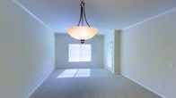 This single-floor space presents a clean, well-maintained interior highlighted by a spacious main room visible in the image. The room features soft, neutral-toned wall-to-wall carpeting that contributes to a calm and comfortable atmosphere. Crown molding lines the upper perimeter of the walls, adding a subtle architectural detail that elevates the overall finish of the space.

A prominent bowl-style pendant light fixture with a dark bronze frame hangs centrally from the ceiling, serving as a functional and decorative focal point. The ceiling height appears standard, providing an open and airy feel throughout the room.

Natural light enters generously through two windows fitted with white horizontal blinds, casting soft patches of sunlight onto the carpeted floor. A door with a glass panel is also visible, likely leading to an exterior area and further enhancing the room's access to natural light. The walls are painted in a light, neutral tone that complements the overall brightness of the space.

The room's layout offers flexible use of the available floor space, making it adaptable for various furniture arrangements. The overall ambiance is clean, understated, and move-in ready.