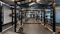 Located at 870 Toftrees Ave in State College, Pennsylvania, this single-floor fitness center is a comprehensively outfitted facility designed to support a wide range of workout routines. The space is finished with durable rubber flooring throughout and features bold blue accent walls paired with neutral grey tones, creating a focused and energizing atmosphere.

The cardio area is anchored by a row of Matrix treadmills and elliptical machines positioned along a wall of large windows, allowing natural light to fill the space and providing views to the outdoors. Pendant and linear LED lighting fixtures illuminate the facility with a clean, modern aesthetic.

A dedicated strength training zone houses a prominent Matrix multi-functional power rack system, complete with cable attachments, battle ropes, and barbells with plate weights. Full-length mirrors line the walls in this area, enhancing the sense of space and aiding in form correction during lifts.

The selectorized machine section features a variety of Matrix cable and resistance machines suited for targeted muscle training, accompanied by adjustable benches. A tiered dumbbell rack offers a wide selection of free weights, with open floor space and flat benches nearby for versatile use. Safety equipment including a defibrillator and fire extinguisher is visibly accessible. The facility is well-organized, fully equipped, and thoughtfully designed for an efficient and complete workout experience.