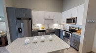 This contemporary single-floor residence showcases a thoughtfully designed open layout with modern finishes throughout. The kitchen is a standout feature, boasting a two-tone cabinetry scheme with crisp white upper cabinets and slate-gray lower cabinets, all complemented by granite-look countertops and a white subway tile backsplash. A large island with matching countertop provides additional workspace and casual seating. Stainless steel appliances, including a refrigerator, dishwasher, over-the-range microwave, and electric range, complete the polished look. Wood-look flooring runs through the kitchen and living areas, adding warmth and continuity. The bedroom is a serene retreat featuring plush carpeting, a ceiling fan, and generously sized windows that invite abundant natural light and offer views of surrounding greenery. The full bathroom includes a soaking tub with shower combination, wood-look flooring, a granite-topped vanity, and clean white cabinetry. The living area is bright and welcoming, enhanced by tall ceilings and large windows that bathe the space in natural light. A cozy seating nook with a pendant light adds character and charm. The overall ambiance is contemporary and sophisticated, with a neutral gray and white palette creating a calm, cohesive atmosphere throughout.