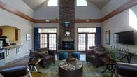 This single-story space makes a striking impression through its dramatic vaulted ceilings that rise to considerable heights, creating an expansive, airy atmosphere. The focal point of the room is a floor-to-ceiling stacked stone fireplace, which anchors the space with a bold, rustic elegance. Flanking the fireplace on both sides are large, multi-pane windows and French doors dressed with deep teal drapery and warm wood trim, allowing generous natural light to fill the interior throughout the day.

The seating area is thoughtfully arranged with dark brown leather armchairs positioned symmetrically around a round, upholstered ottoman serving as a central coffee table. Teal fabric ottomans provide additional accent seating and tie together the color palette of the room. A patterned area rug in earthy greens and blues grounds the conversation area and adds a layer of visual interest to the space.

A warm wood trim band runs along the upper perimeter of the walls, bridging the transition between the lower wall sections and the soaring ceiling above. The overall ambiance is welcoming and sophisticated, blending rustic and contemporary design elements seamlessly.