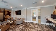Located in Greenville, SC, this single-floor community amenity space offers a well-appointed and contemporary environment designed for both collaboration and relaxation. The layout is thoughtfully organized into distinct zones, each serving a specific purpose while maintaining a cohesive modern aesthetic throughout.

A standout feature is the private meeting room, enclosed with large glass partition walls that allow natural light to flow through while maintaining a sense of privacy. Inside, a solid dark wood conference table is surrounded by vibrant mustard-yellow upholstered chairs, anchored by a bold oversized pendant light overhead. A dramatic large-scale photographic mural serves as a striking accent wall, adding depth and visual interest to the space.

The open lounge areas are furnished with comfortable seating arrangements, including plush gray sofas and deep burgundy accent chairs paired with compact side tables — ideal for casual gatherings or informal meetings. Wall-mounted flat-screen televisions are positioned throughout the lounge for entertainment and presentations. The space is tied together by richly textured, patterned carpet flooring in earthy tones that adds warmth and character underfoot.

An open-railing overlook introduces an architectural element that enhances the sense of openness, while recessed lighting and large windows ensure the space remains bright and welcoming. The overall ambiance is polished, functional, and inviting.